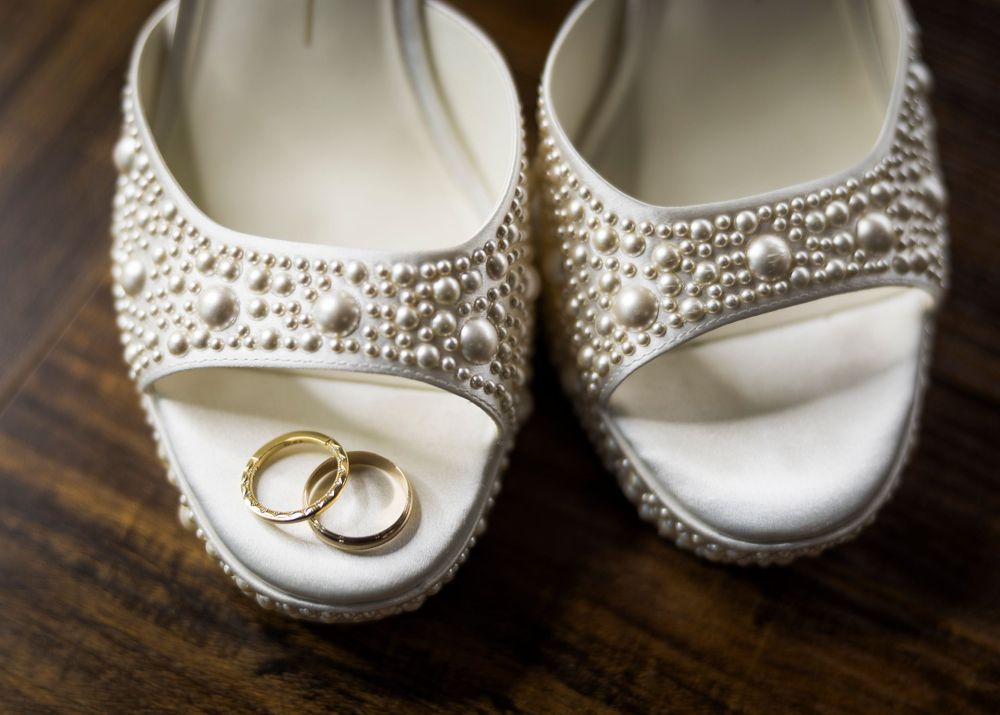 Pearl-embellished bridal heels with two gold wedding rings resting between them on a wooden surface.