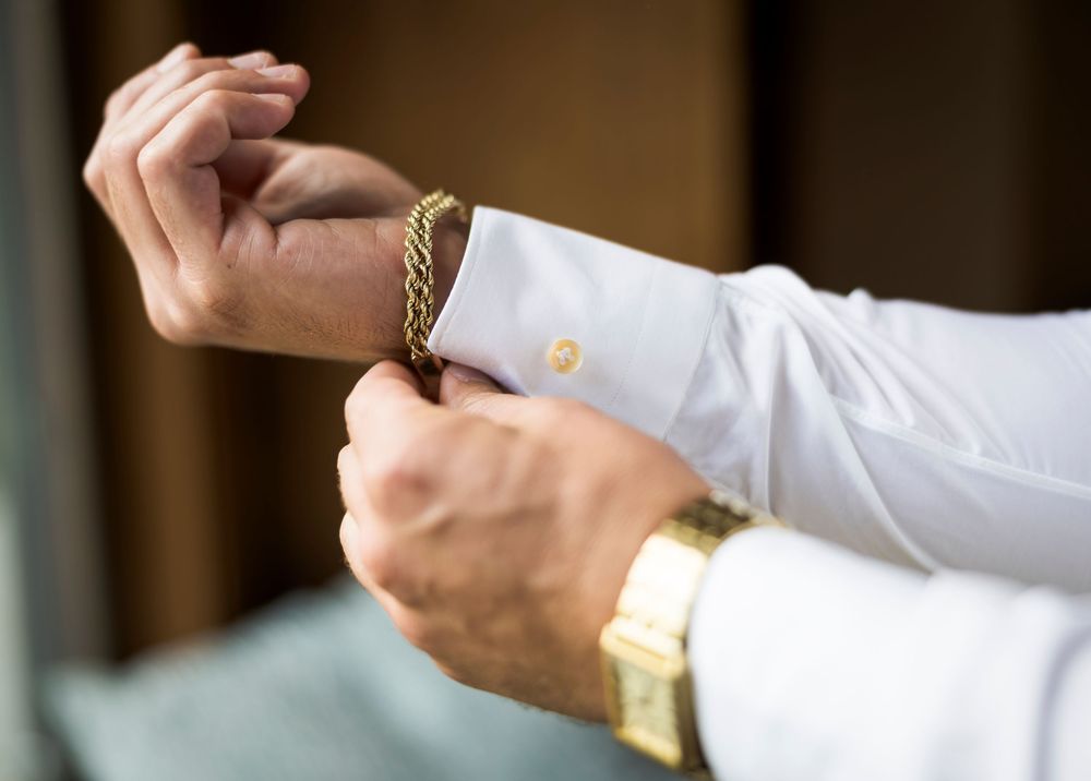 Groom fastening a gold chain bracelet on his wrist while wearing a white dress shirt and gold watch.