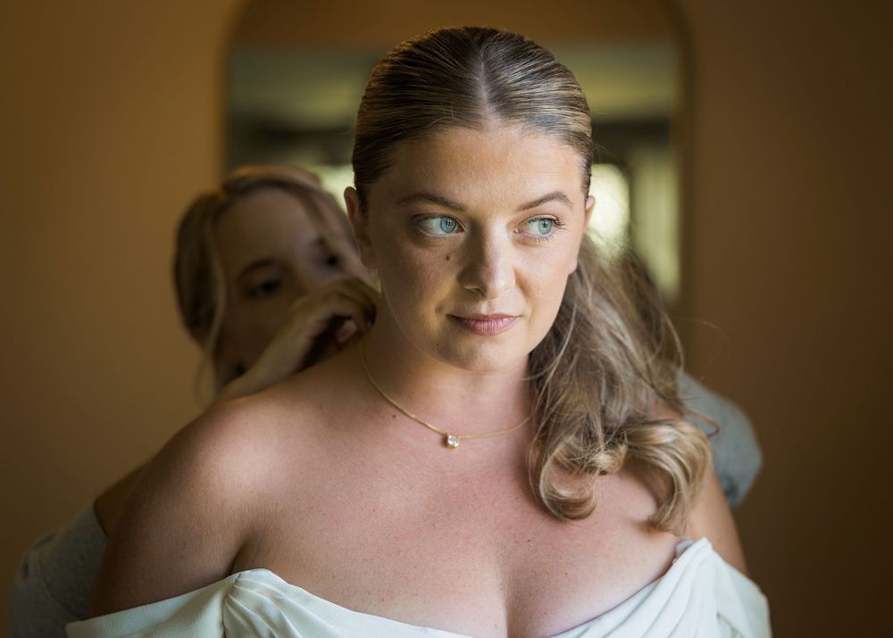 Bride in white dress getting ready with bridesmaid assistance, wearing delicate necklace.