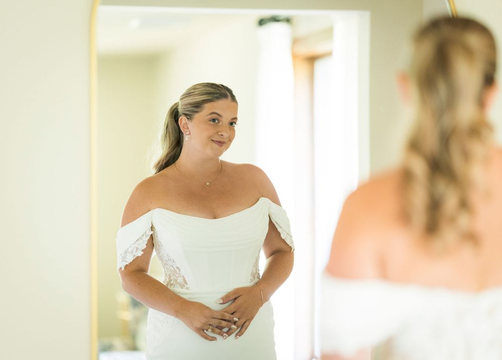 Elegant bride in off-shoulder white lace wedding gown admires herself in mirror before ceremony.