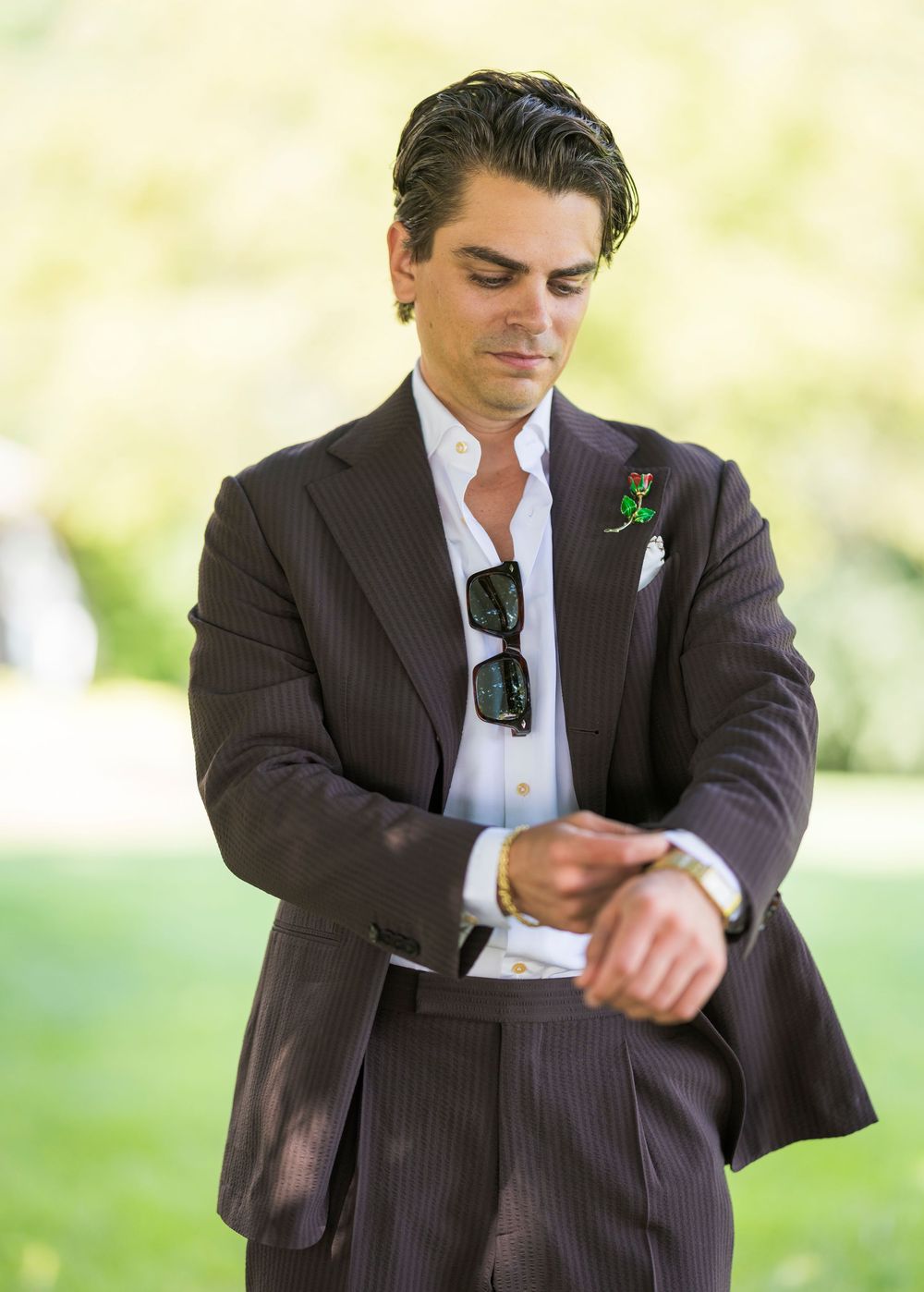 Stylish groom in dark pinstripe suit checking his watch with sunglasses tucked in pocket and green boutonniere.