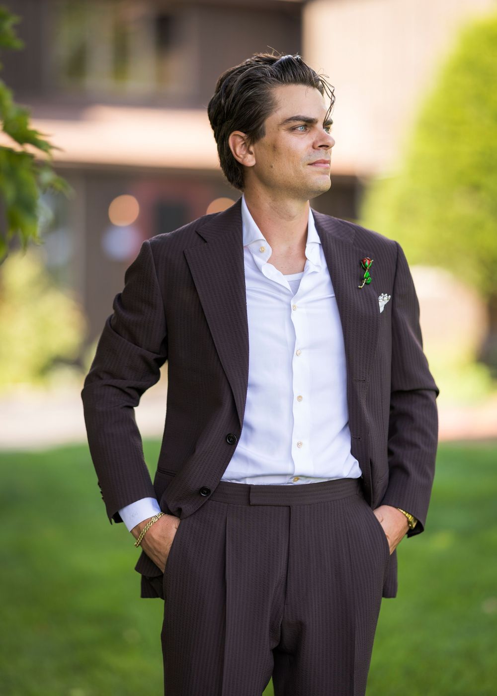 Elegant groom in dark pinstripe suit with green boutonniere and white pocket square standing outdoors.