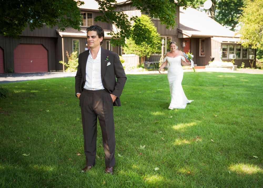 Groom in dark suit stands on green lawn as bride in white gown approaches from behind at outdoor wedding venue.