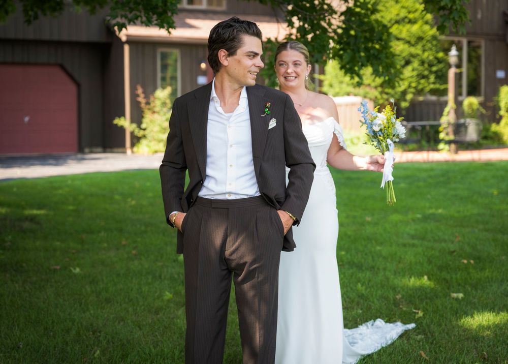 Bride and groom posing outdoors on green lawn, groom in dark suit, bride in elegant white gown.