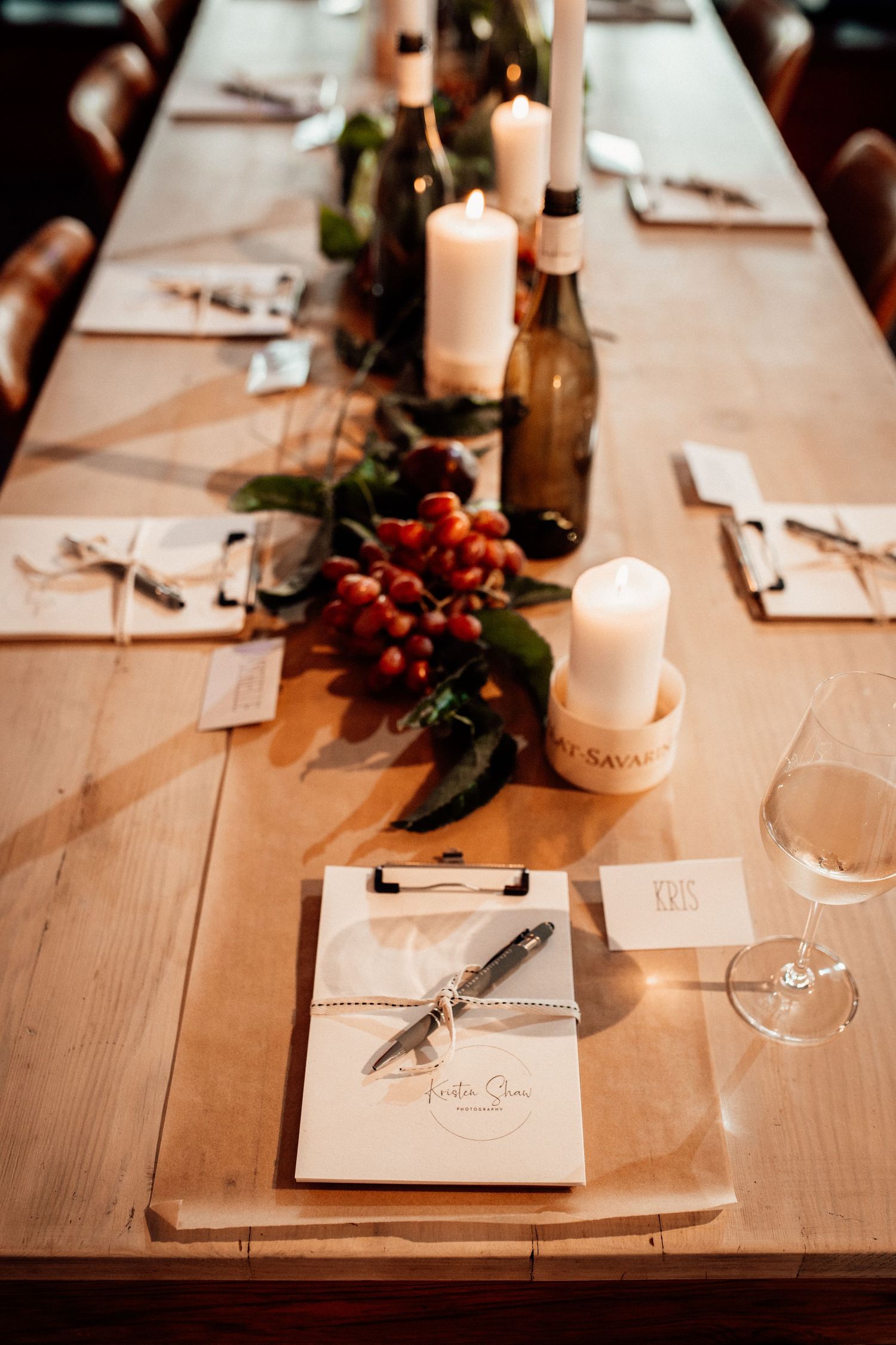 Elegant rustic wedding table setting with white candles, red berries, menus on clipboards, and wine glasses on wooden table.