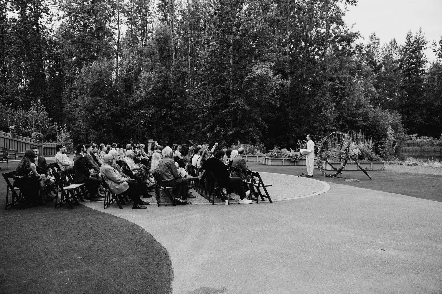 Bride in white gown with fur wrap holds clear umbrella in rainy park, groom stands near windmill during Redmond Wedding