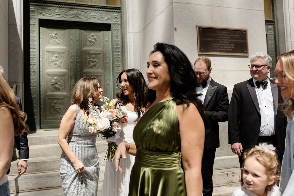 Wedding party on steps, bridesmaid in green satin dress holding bouquet, guests in formal attire outside ornate building.