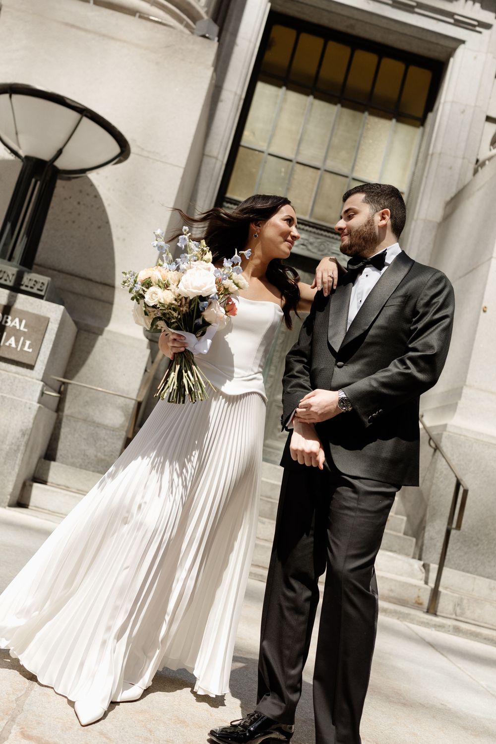 Bride in white pleated gown holding bouquet stands with groom in black tuxedo outside elegant building.