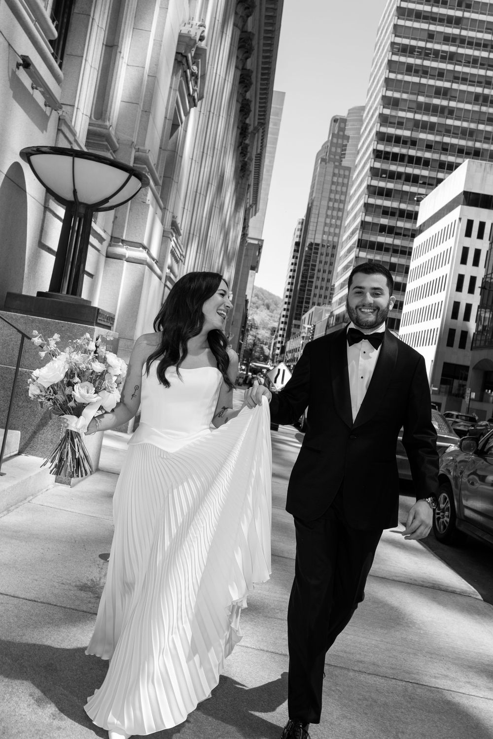 Black and white photo of a smiling bride and groom walking together on a city street, bride holding a bouquet.
