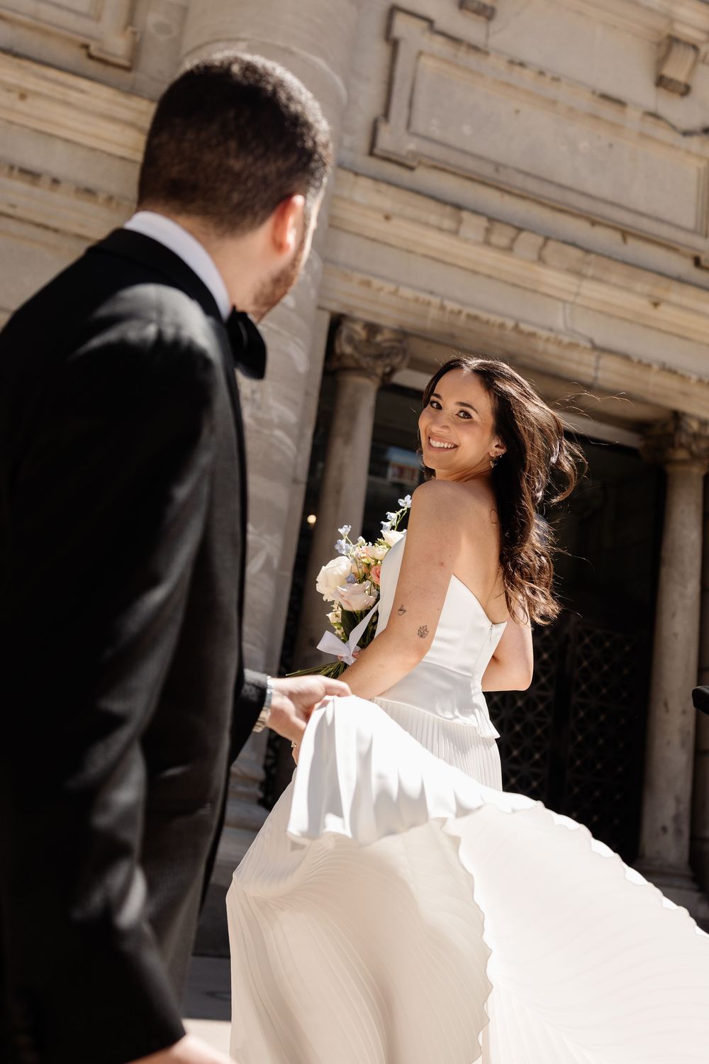 Smiling bride in white gown holds bouquet while groom in black suit leads her by hand near elegant building.