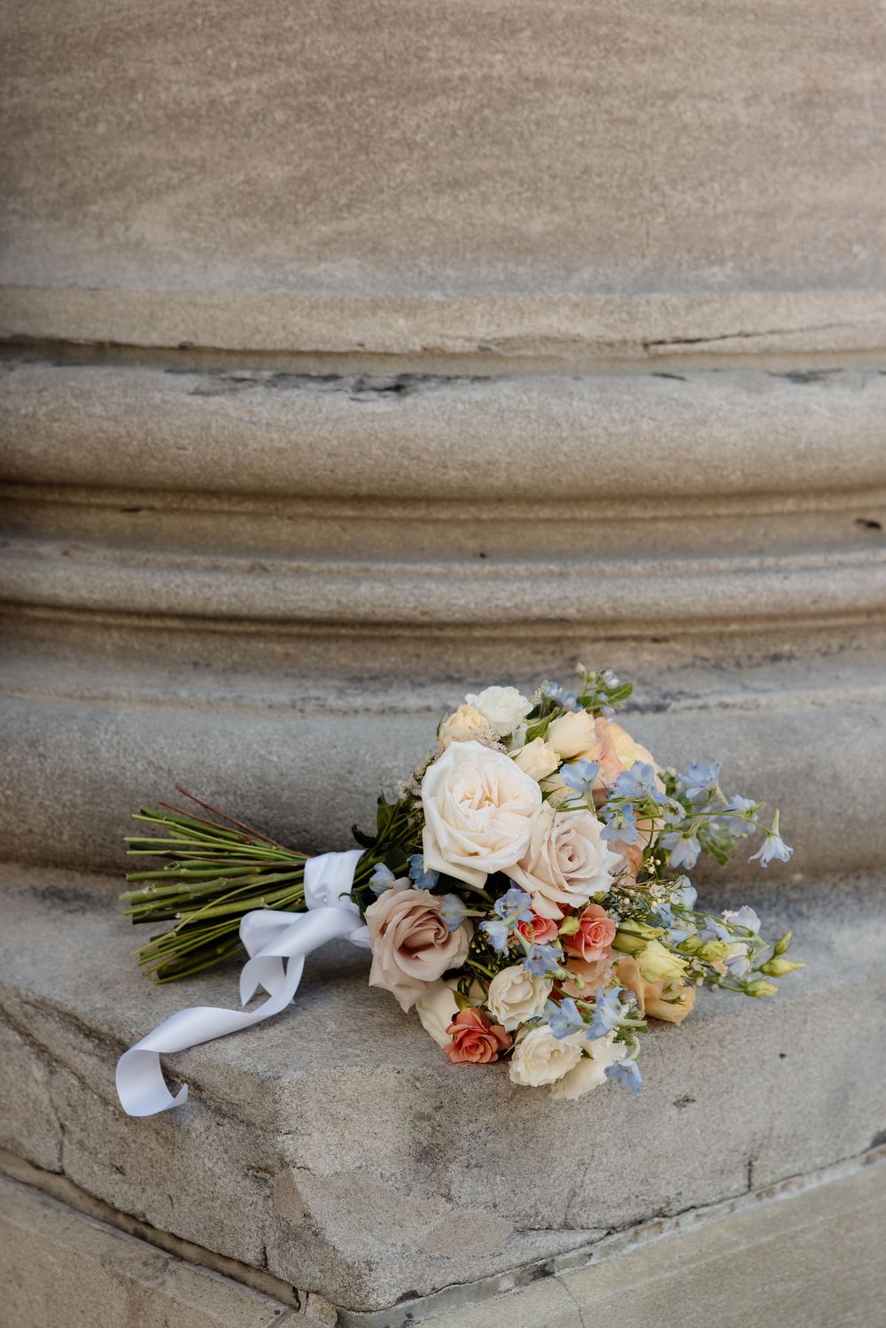 Elegant wedding bouquet with peach roses and blue flowers tied with white ribbon resting on stone steps.