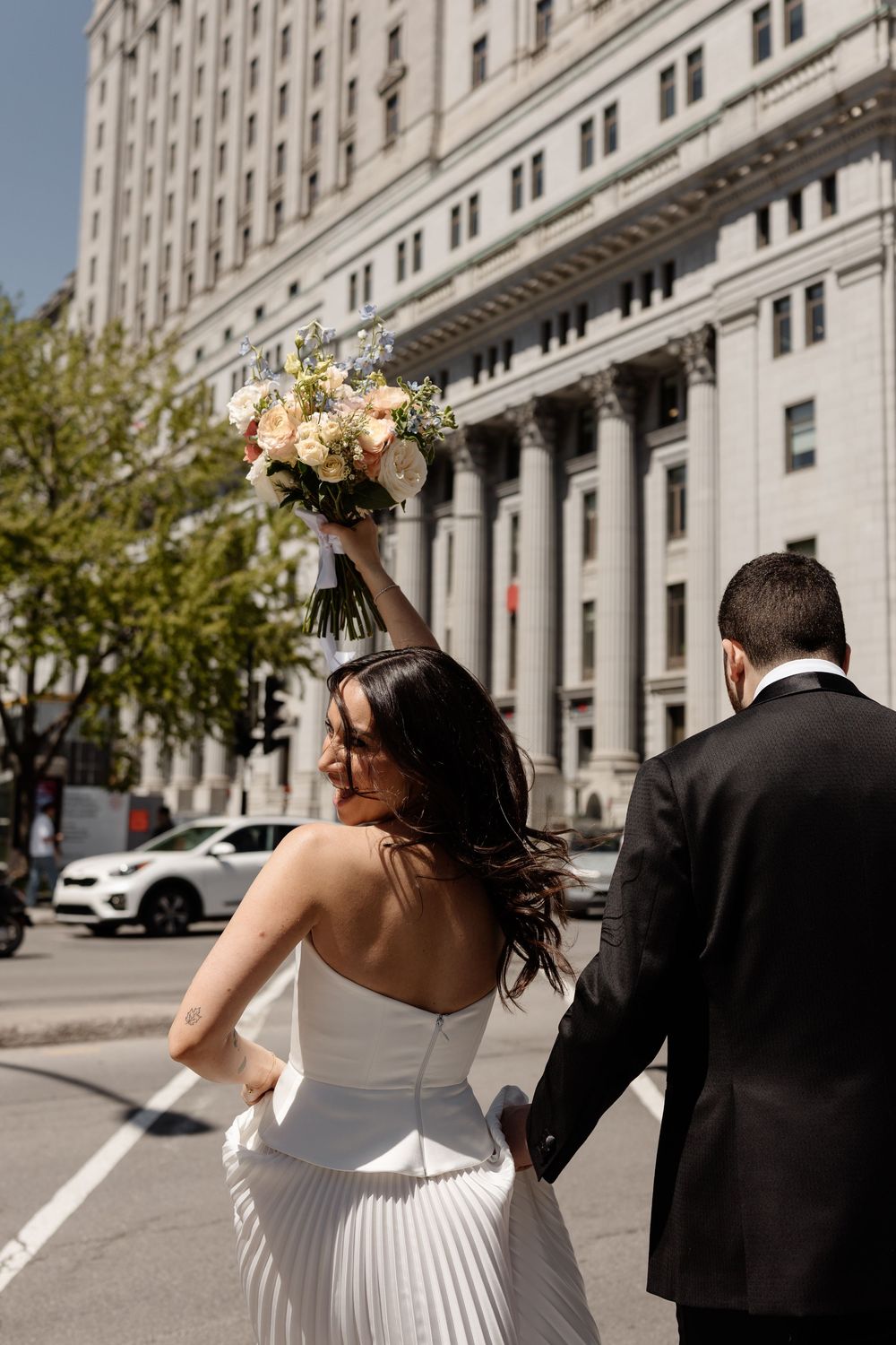 Bride in white gown raises bouquet while walking with groom in black suit past grand building on sunny city street.