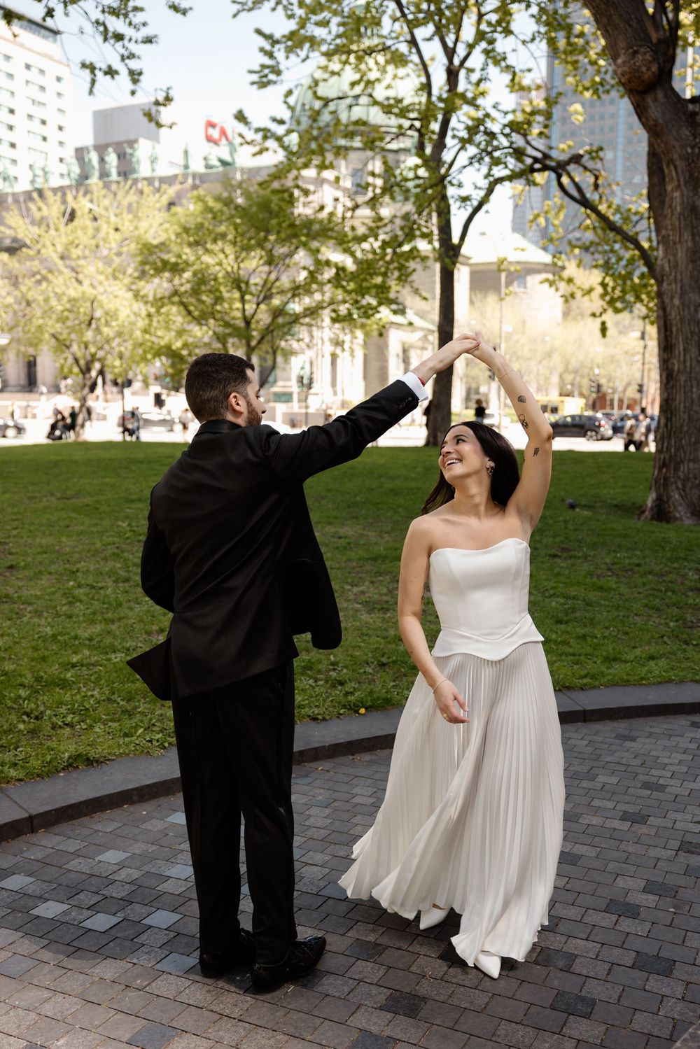 Bride in white strapless gown spins as groom in black suit leads dance in urban park setting.