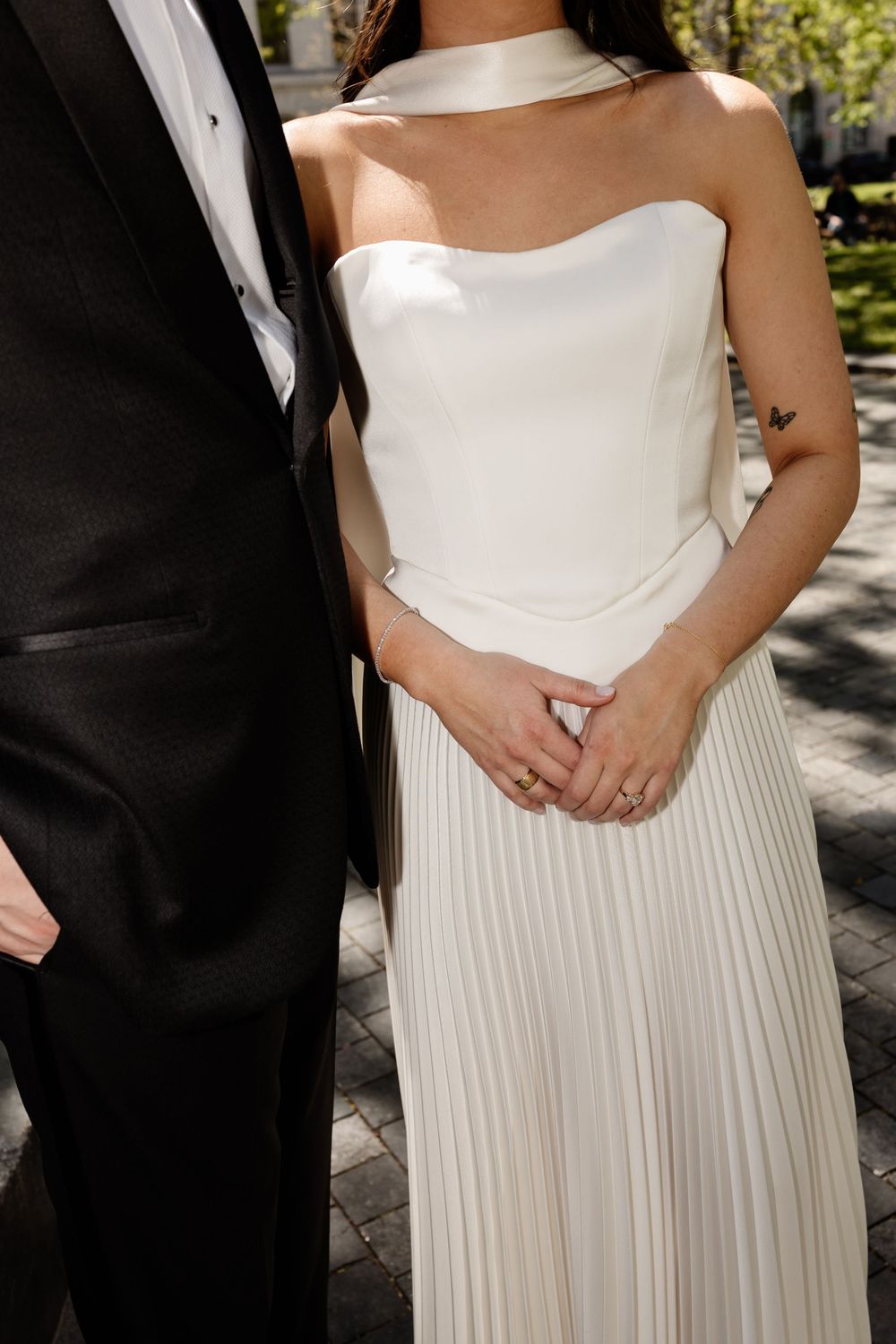 Bride in strapless white pleated gown with choker stands beside groom in black tuxedo outdoors.