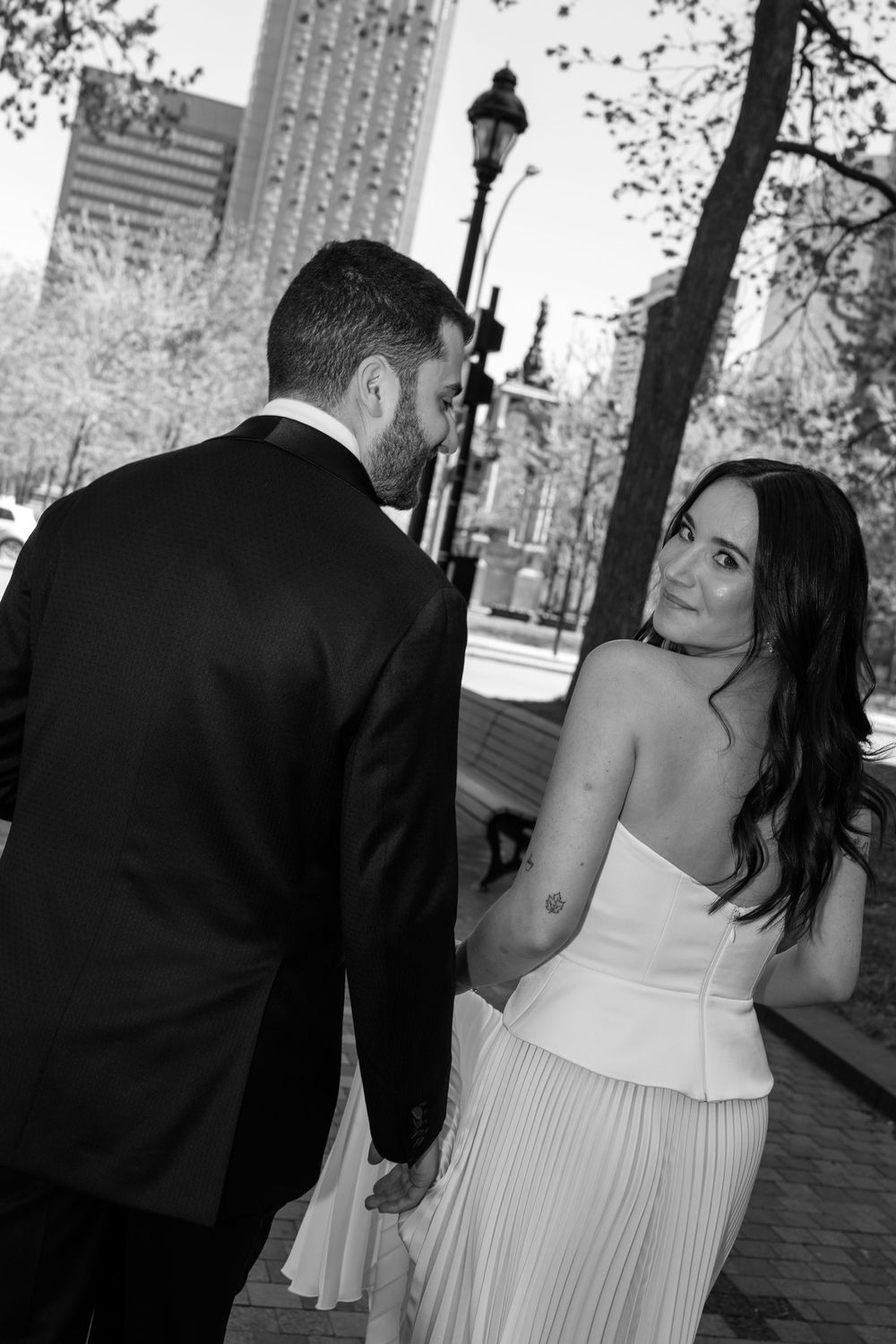 Bride and groom walk hand in hand on city street, bride glancing back in black and white photo.