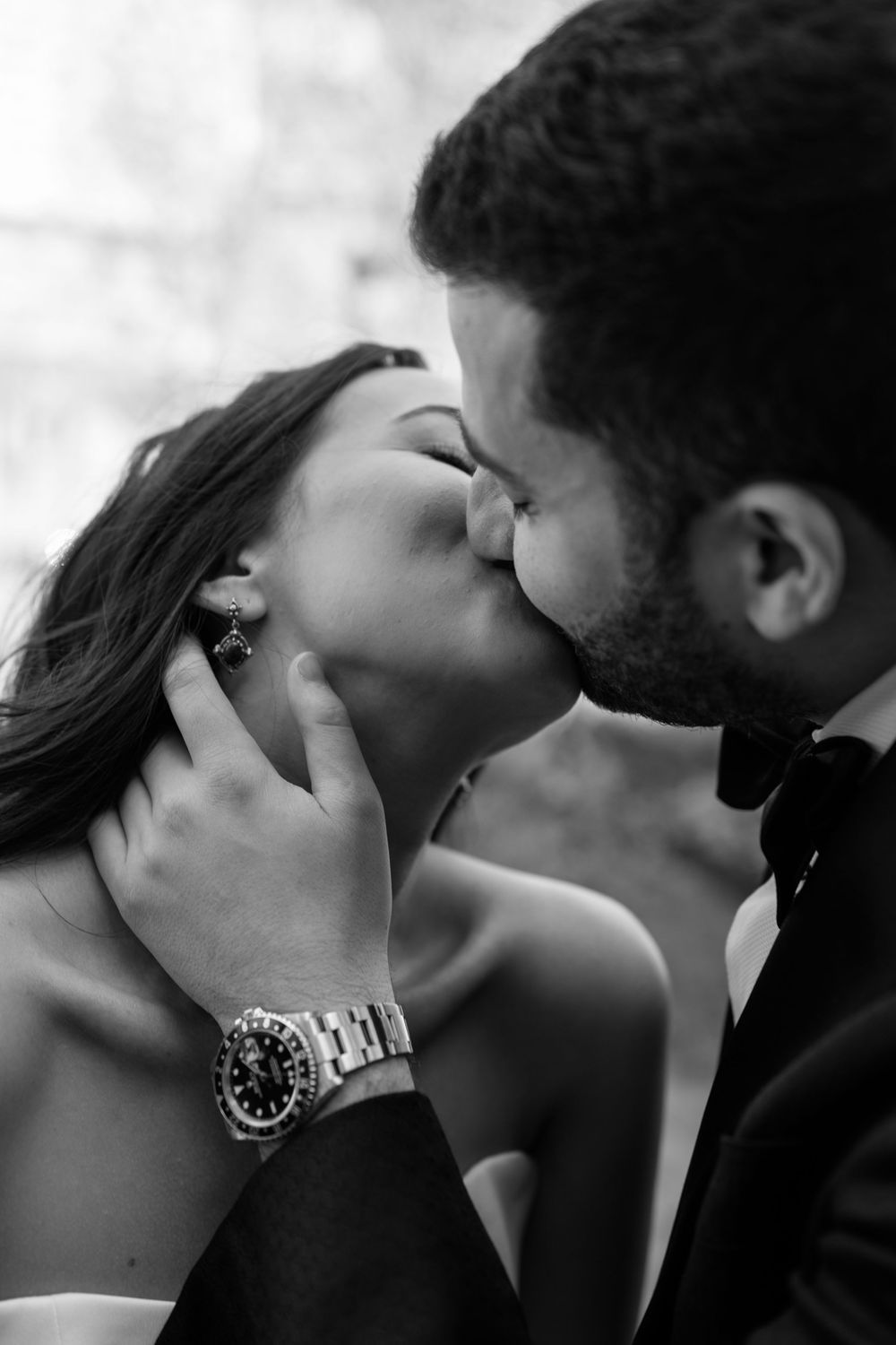 Black and white photo of a couple sharing a romantic kiss, groom in bow tie with Rolex watch visible.