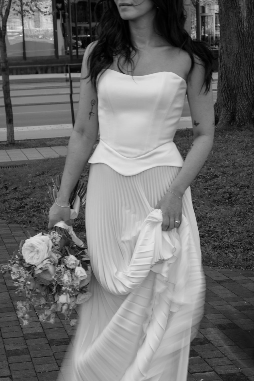 Black and white photo of a bride in a strapless pleated wedding gown holding a floral bouquet outdoors.