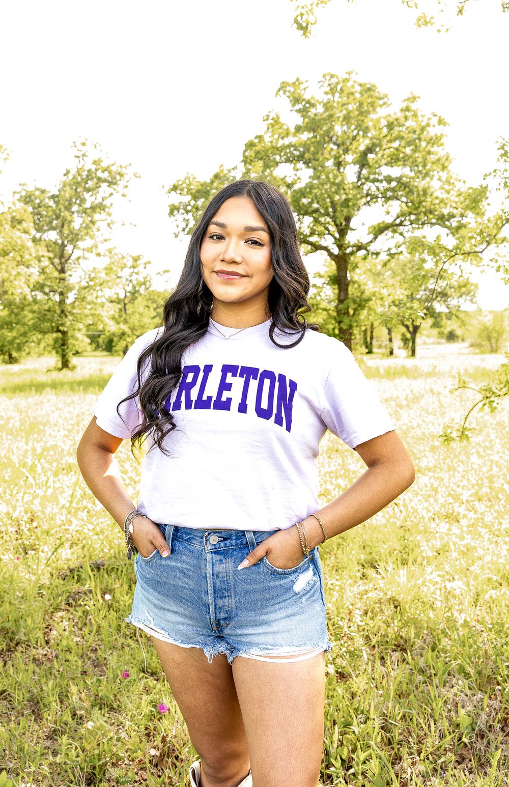 Young woman in Tarleton college shirt and denim shorts stands in a sunny wildflower meadow.