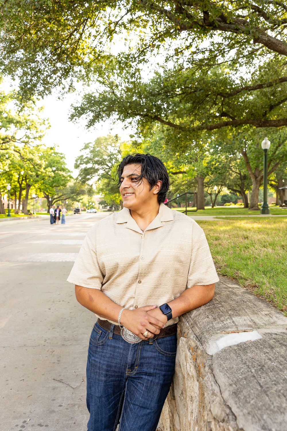 Smiling young man in beige shirt leans against stone wall on tree-lined college campus walkway.