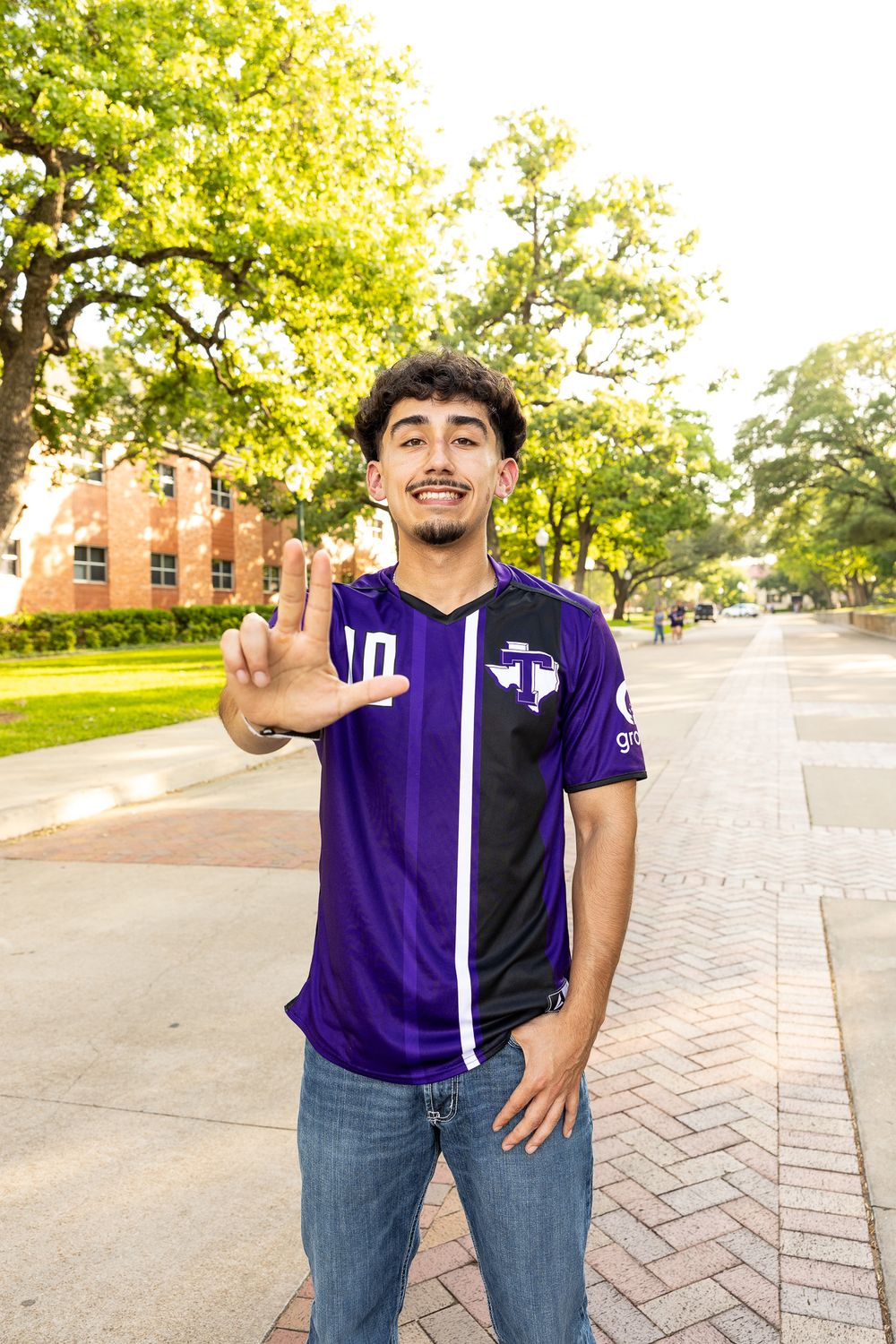 Young man in purple Tarleton jersey makes hand sign on a tree-lined university campus walkway.