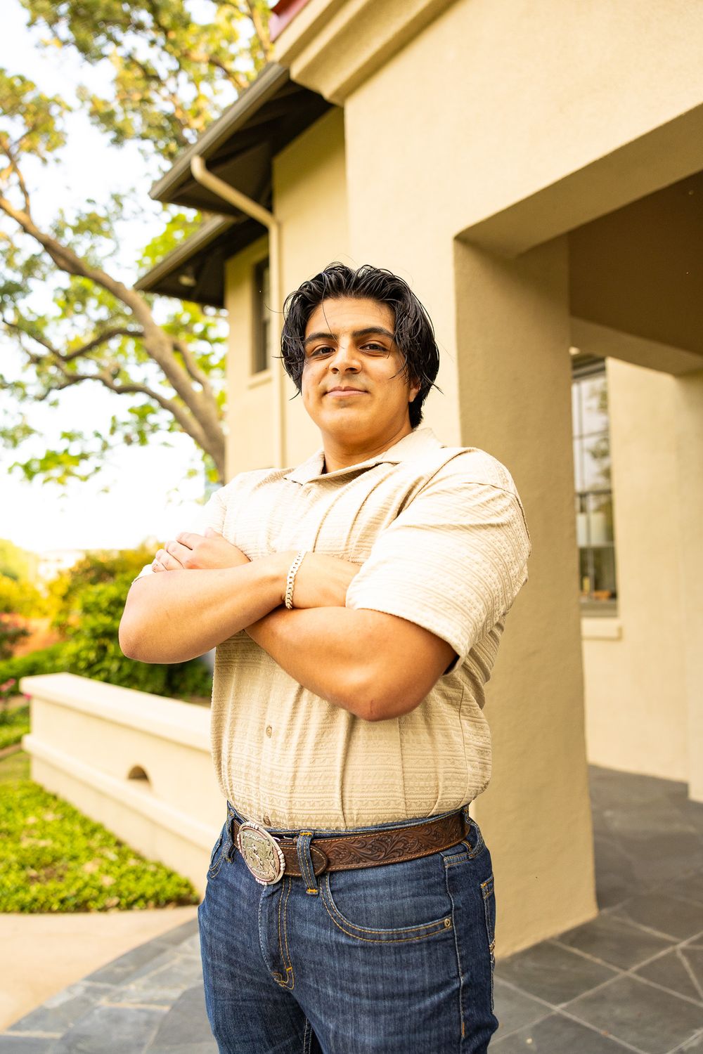 Confident young man in beige shirt stands with arms crossed outside a building in warm sunlight.