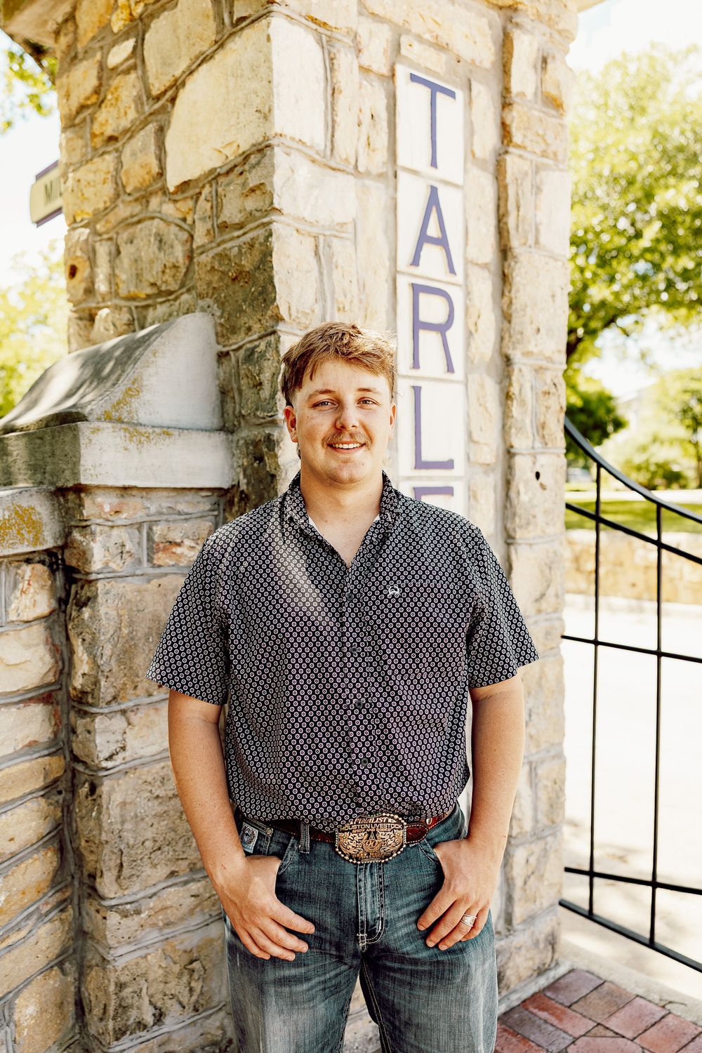 Smiling young man in patterned shirt and jeans poses beside a stone Tarleton State University entrance sign.