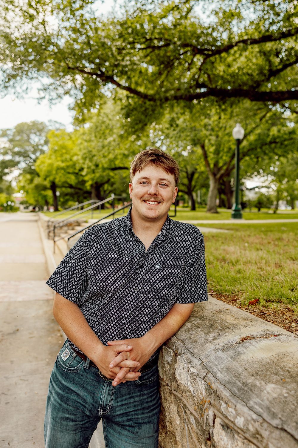 Smiling young man in patterned shirt leans against a stone wall on a sunny college campus pathway.