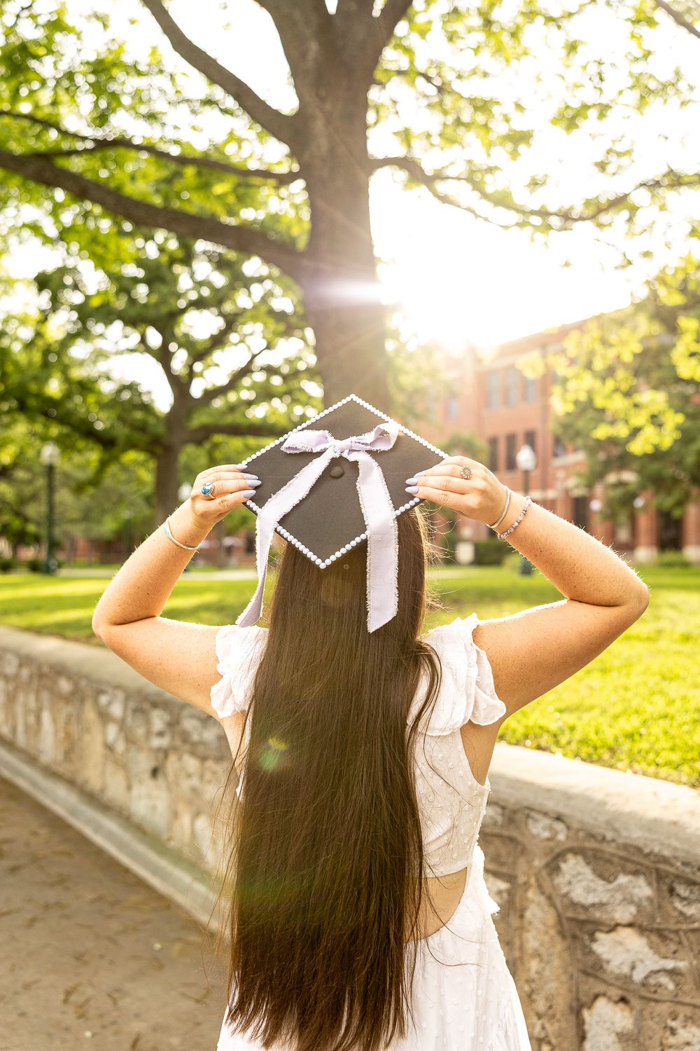 Graduate holds black mortarboard cap with white ribbon in sunlit campus setting, viewed from behind.