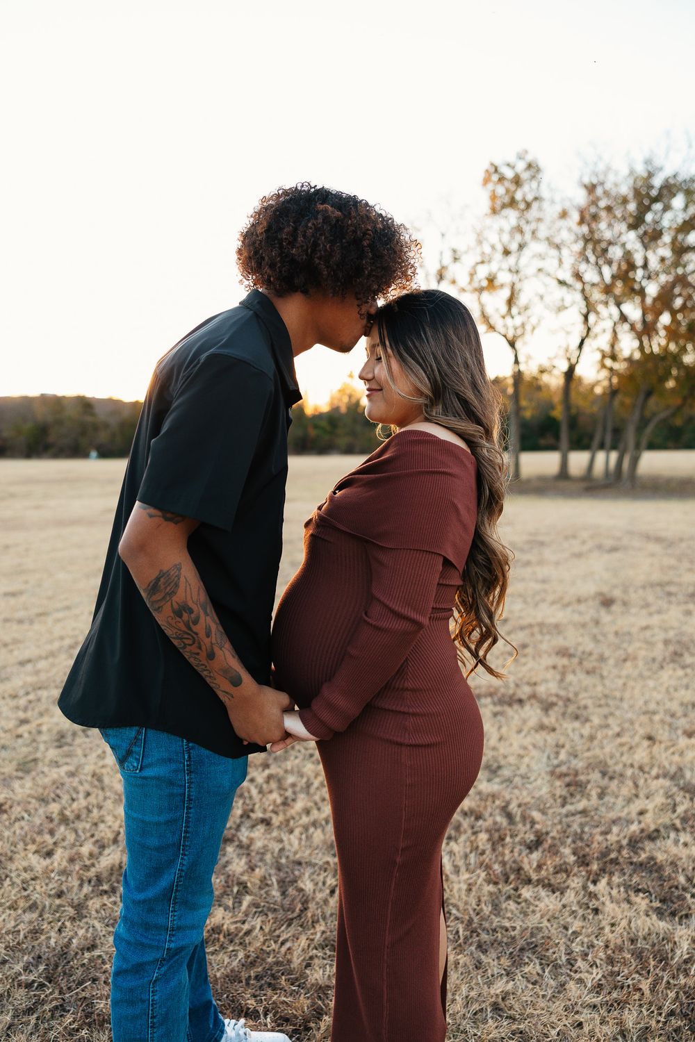 Expecting couple holds hands facing each other in golden sunset field with autumn trees.