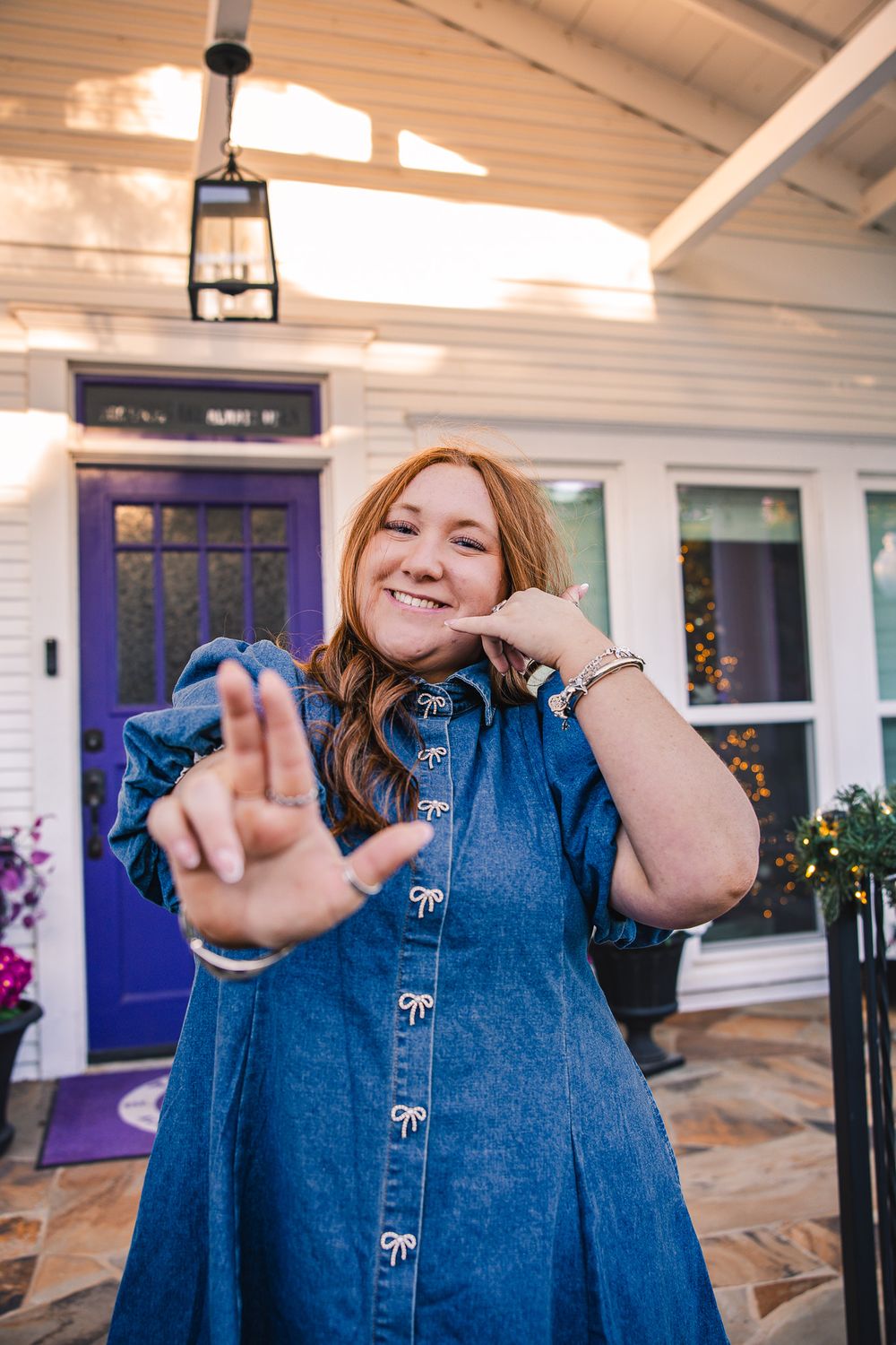 Smiling redhead woman in denim bow dress posing playfully in front of a home with purple door.