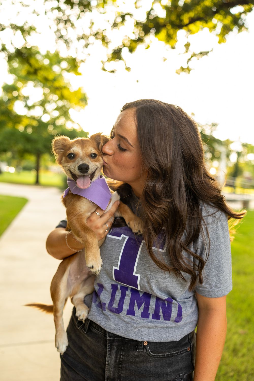 Young woman kissing her small tan dog wearing a purple bandana outdoors in golden light.