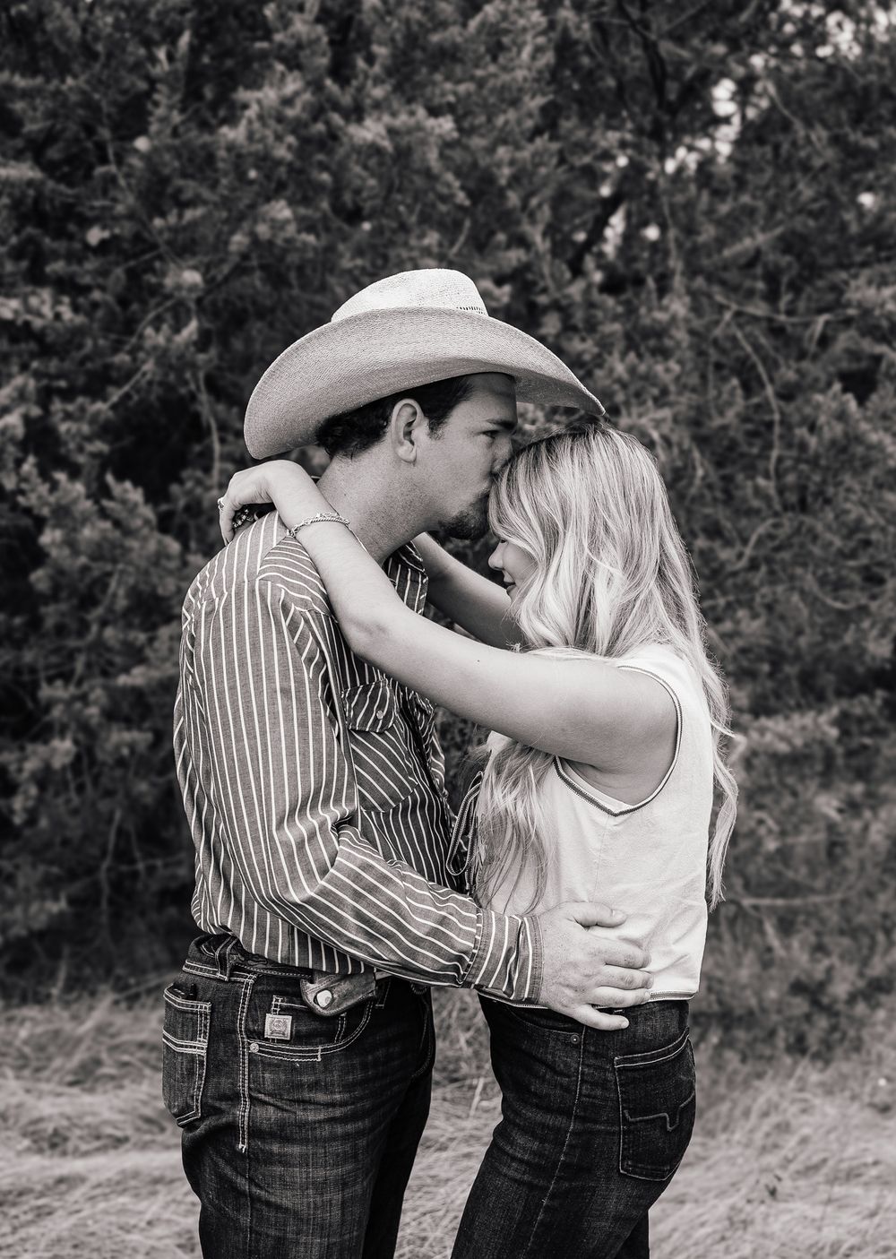 Black and white photo of a young couple sharing a kiss outdoors, man wearing a cowboy hat.