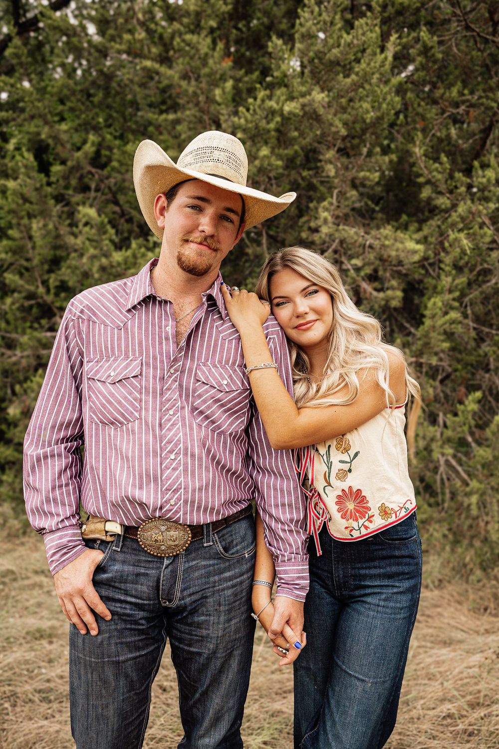 Young couple in western wear pose outdoors, man in cowboy hat and striped shirt, woman in floral top and jeans.