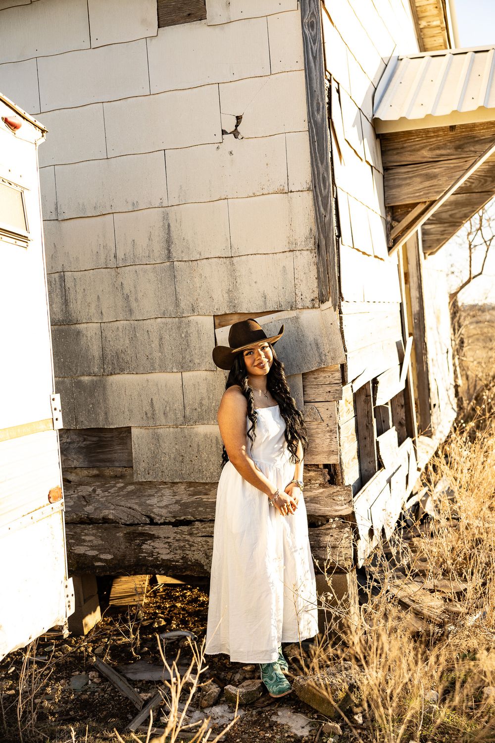 Young woman in white dress and black cowboy hat poses by weathered wooden building in rustic setting.