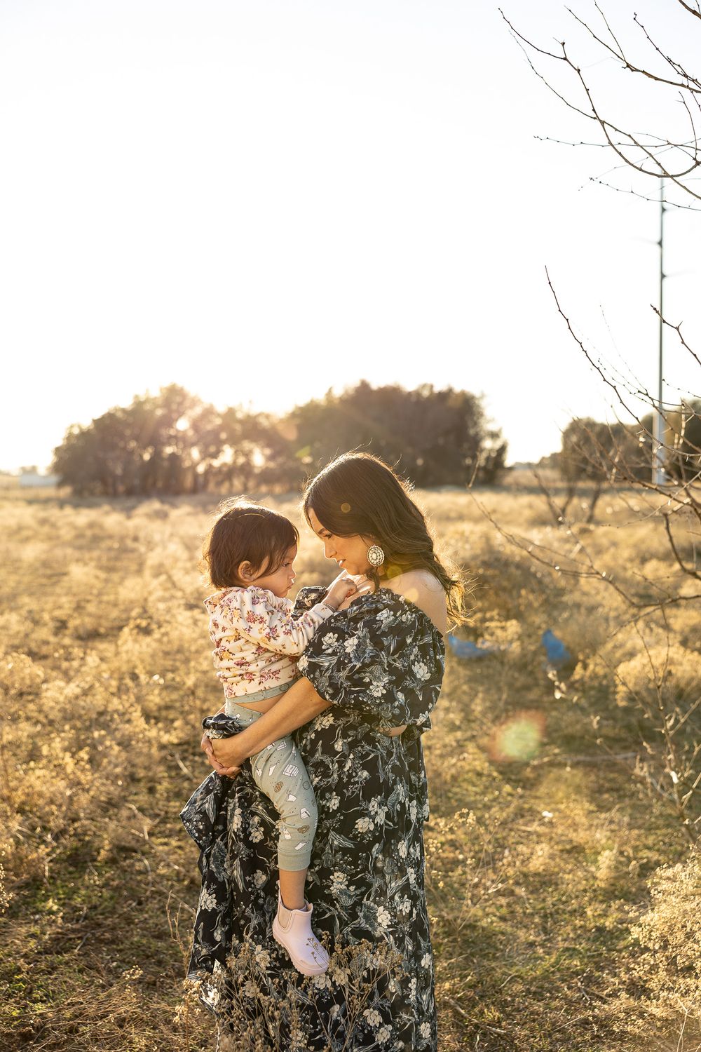 Mother in floral dress holds young toddler in golden sunlit open field with bare trees.