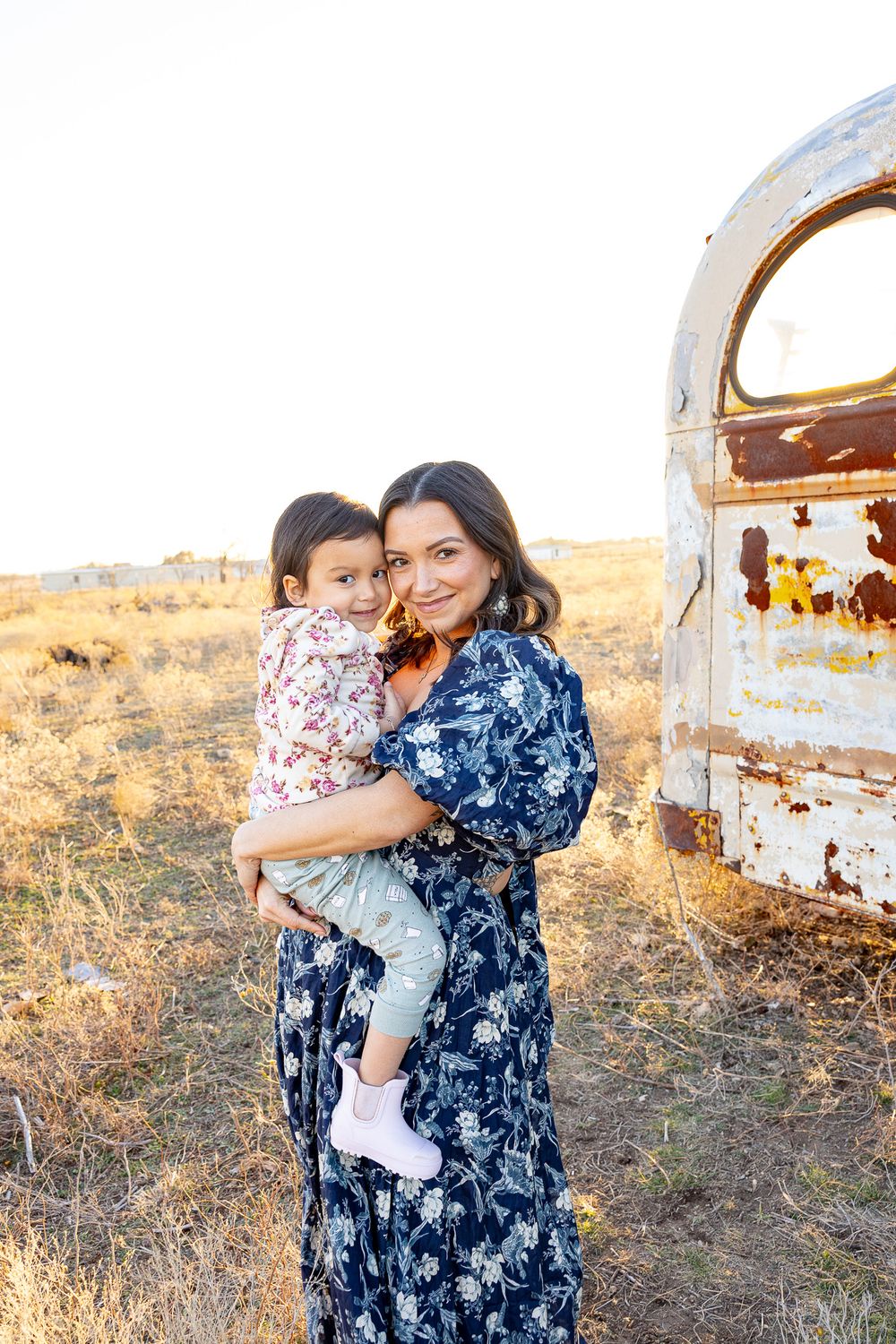 Smiling mother in navy floral dress holds young child near rusty vintage vehicle in dry field.