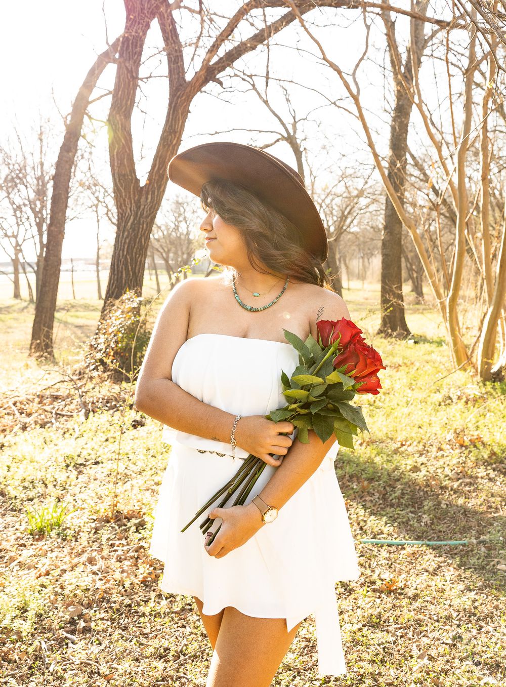 Young woman in white dress and brown hat holds red roses outdoors among bare trees in golden sunlight.