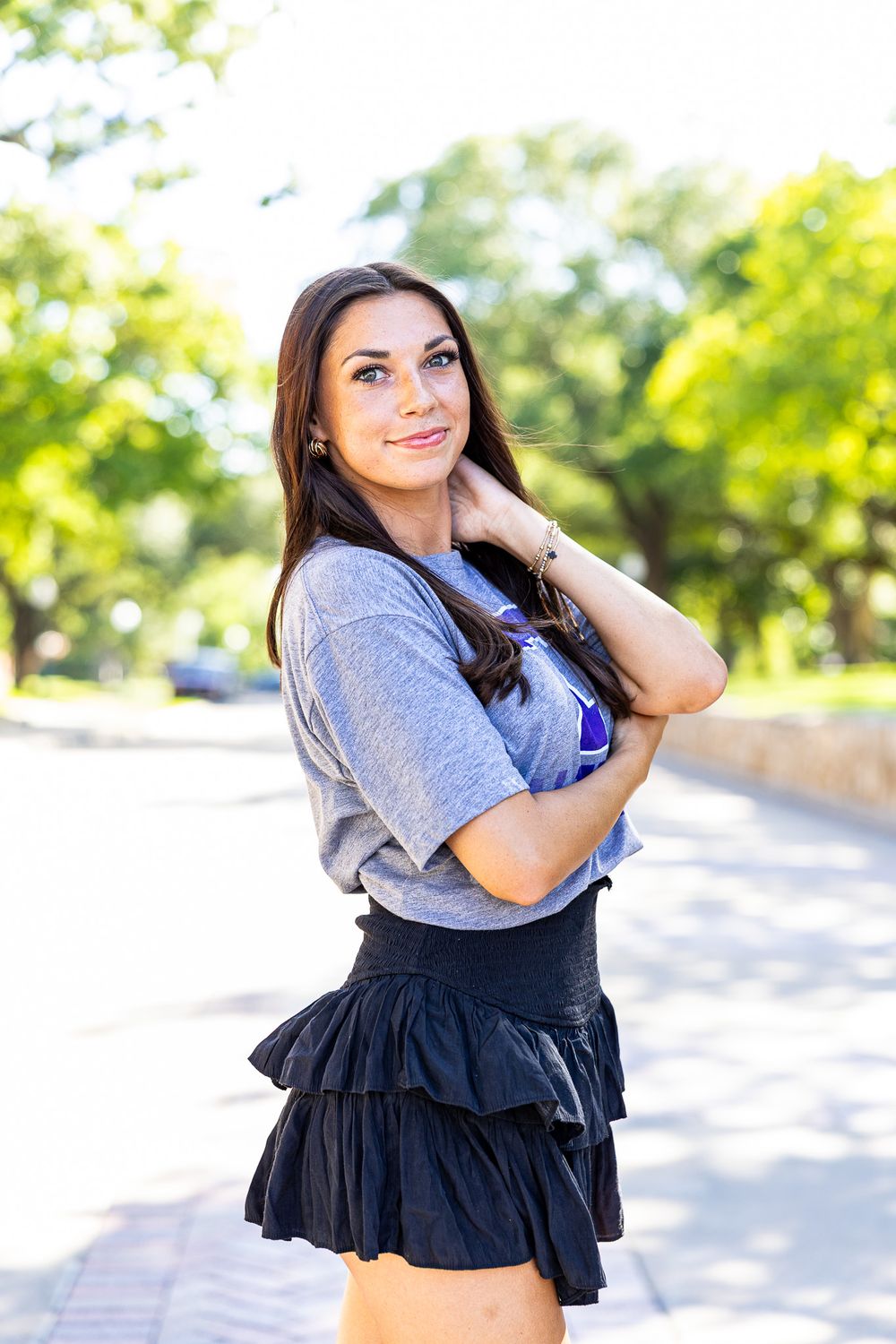 Brunette woman in gray crop top and black skirt poses on sunny tree-lined sidewalk.