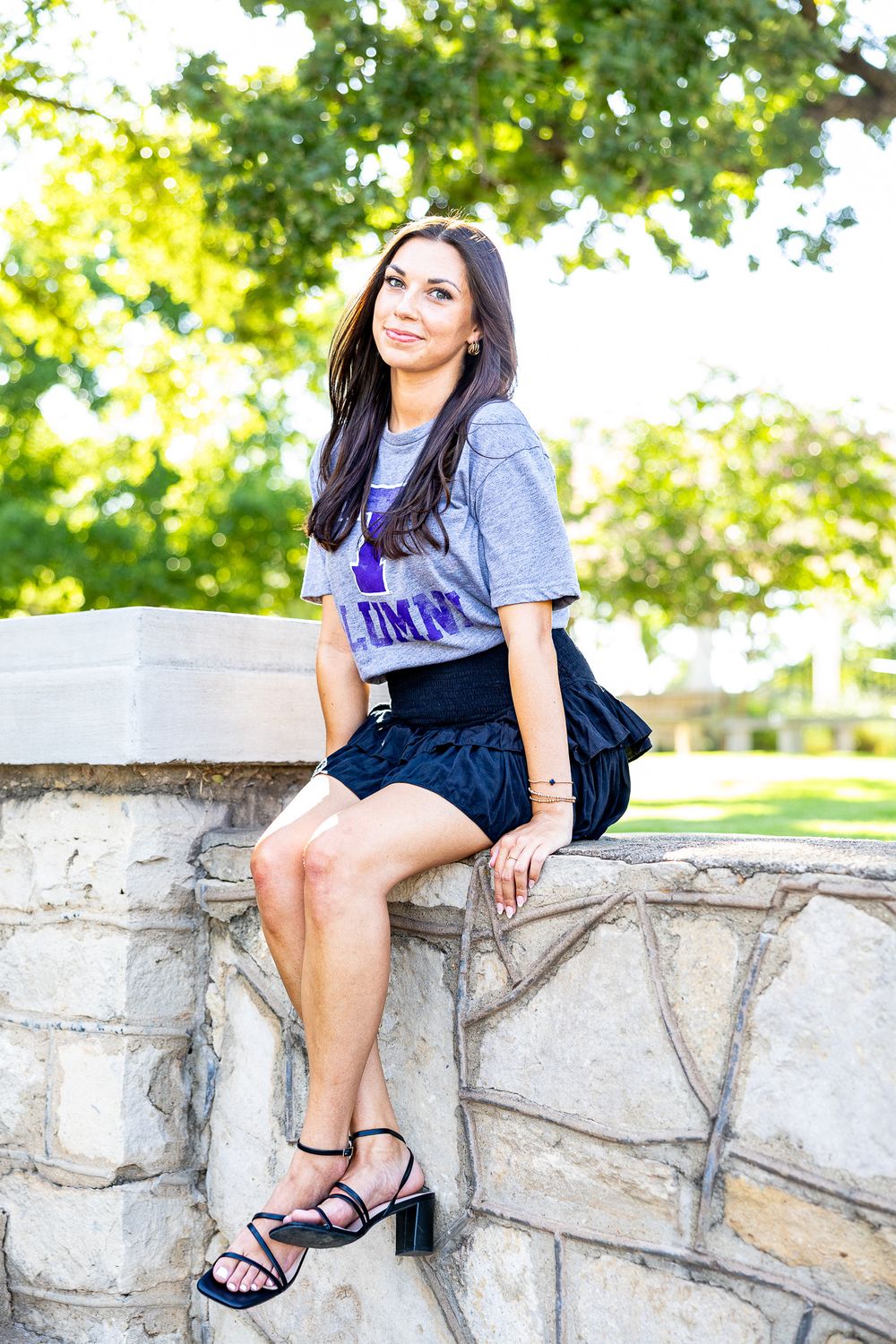 Young woman in purple alumni shirt sits on stone wall in sunny park setting.