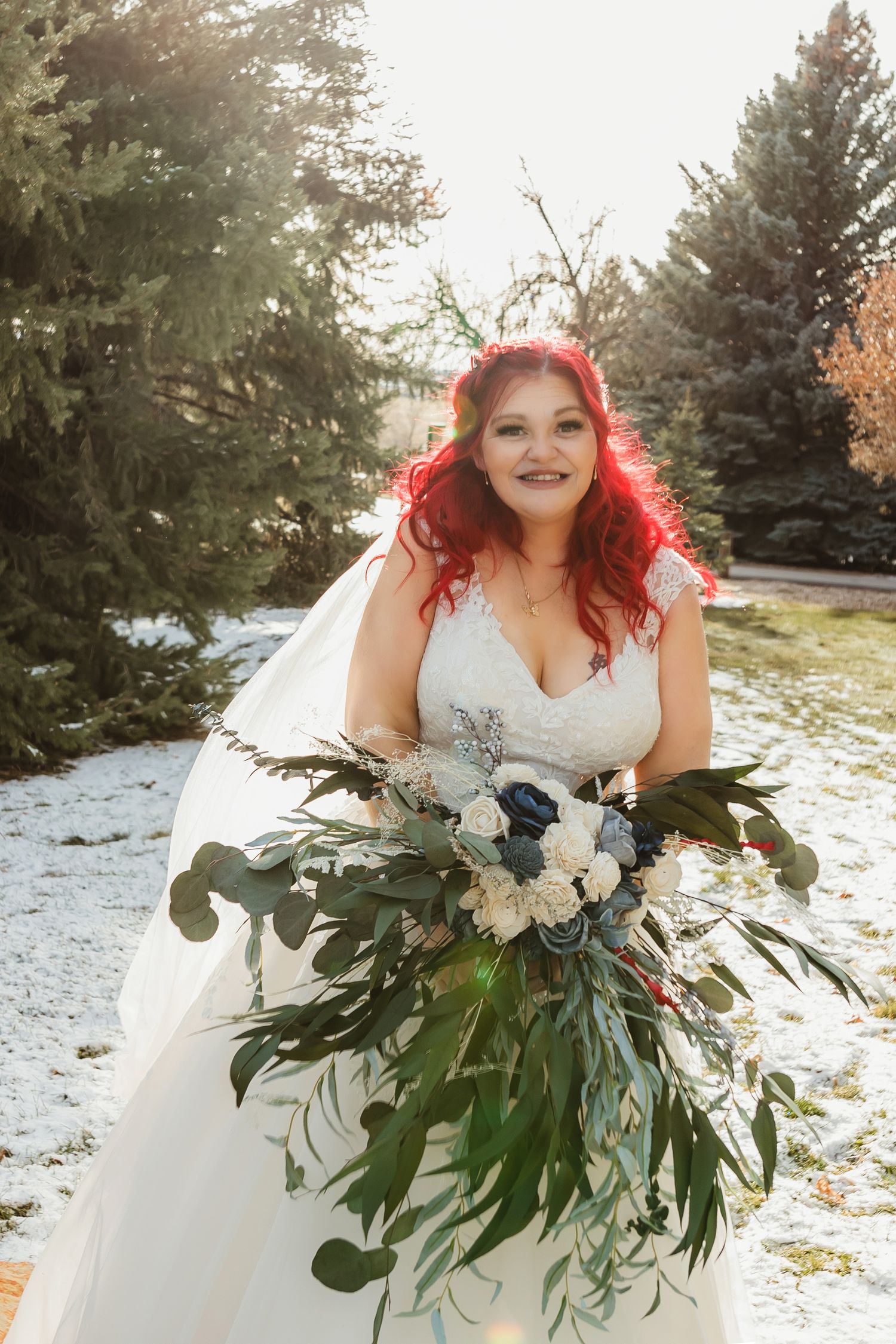 Red-haired bride in white lace gown holds lush greenery bouquet with navy flowers outdoors in snowy winter setting.