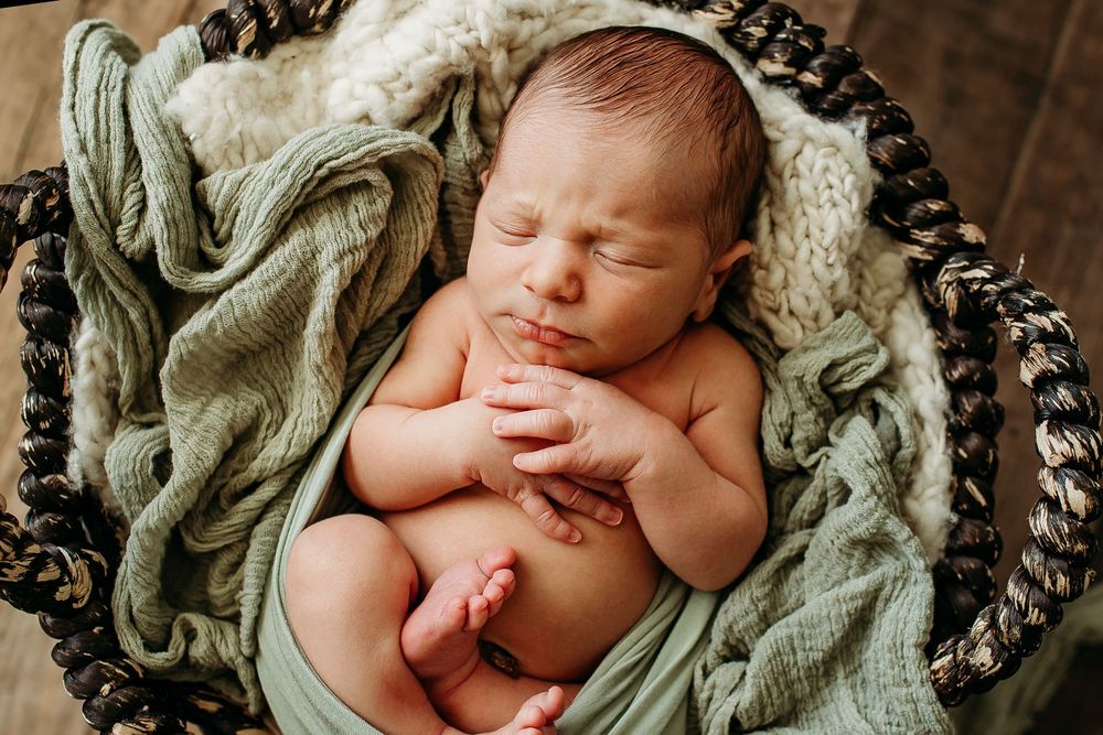 Sleeping newborn baby wrapped in green cloth resting in a woven basket with soft blanket.
