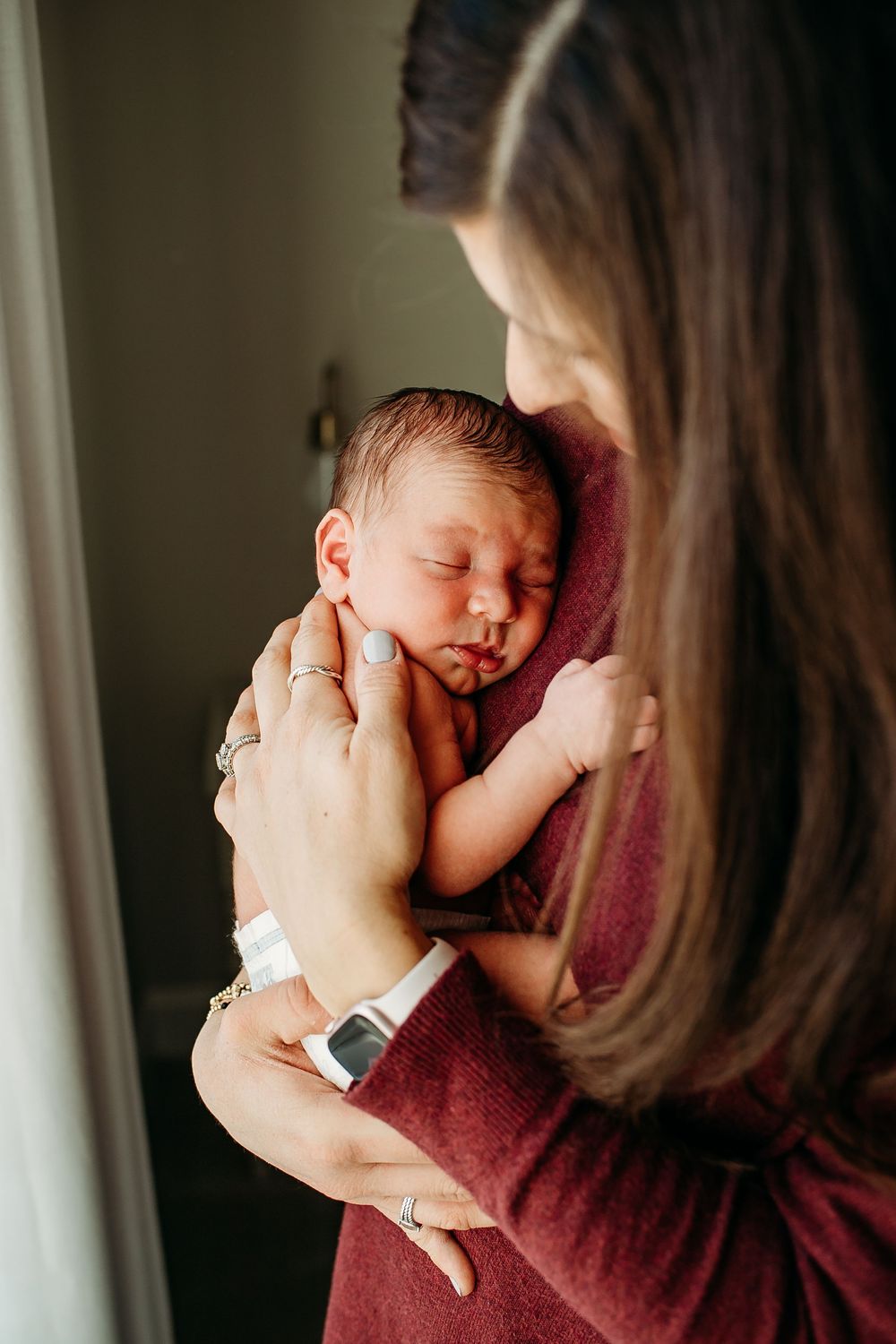 Mother in red sweater tenderly cradling her sleeping newborn baby against her shoulder.