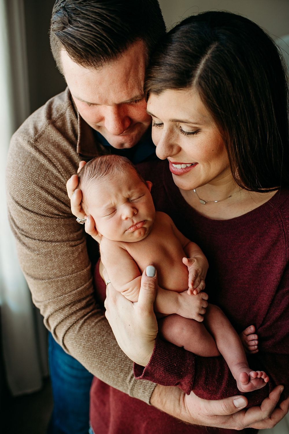 Loving parents in casual attire smiling down at their sleeping newborn baby held between them.