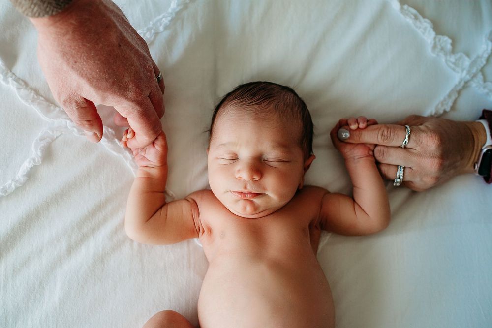 Sleeping newborn baby grasps parents fingers while lying on white blanket.