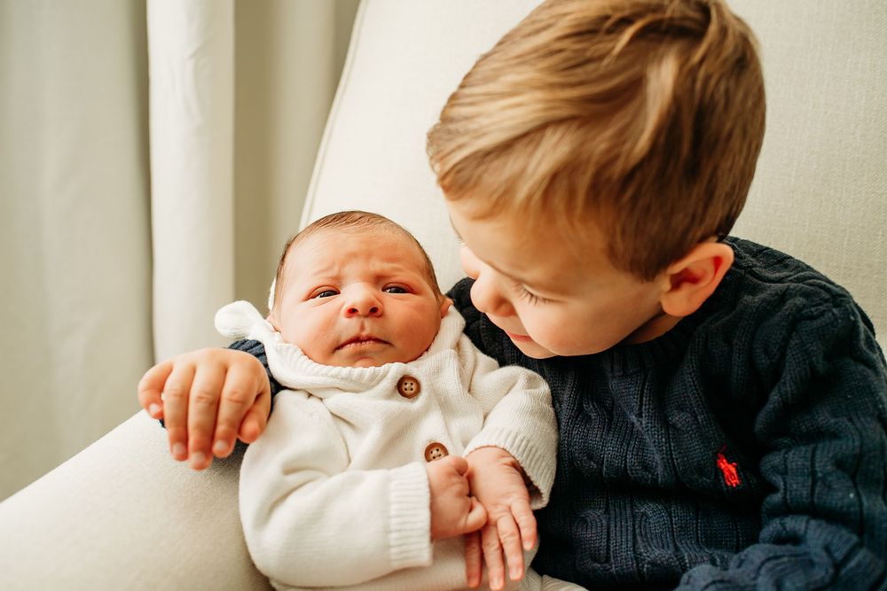 Toddler boy in navy sweater gently holding and looking at his newborn sibling in a white chair.