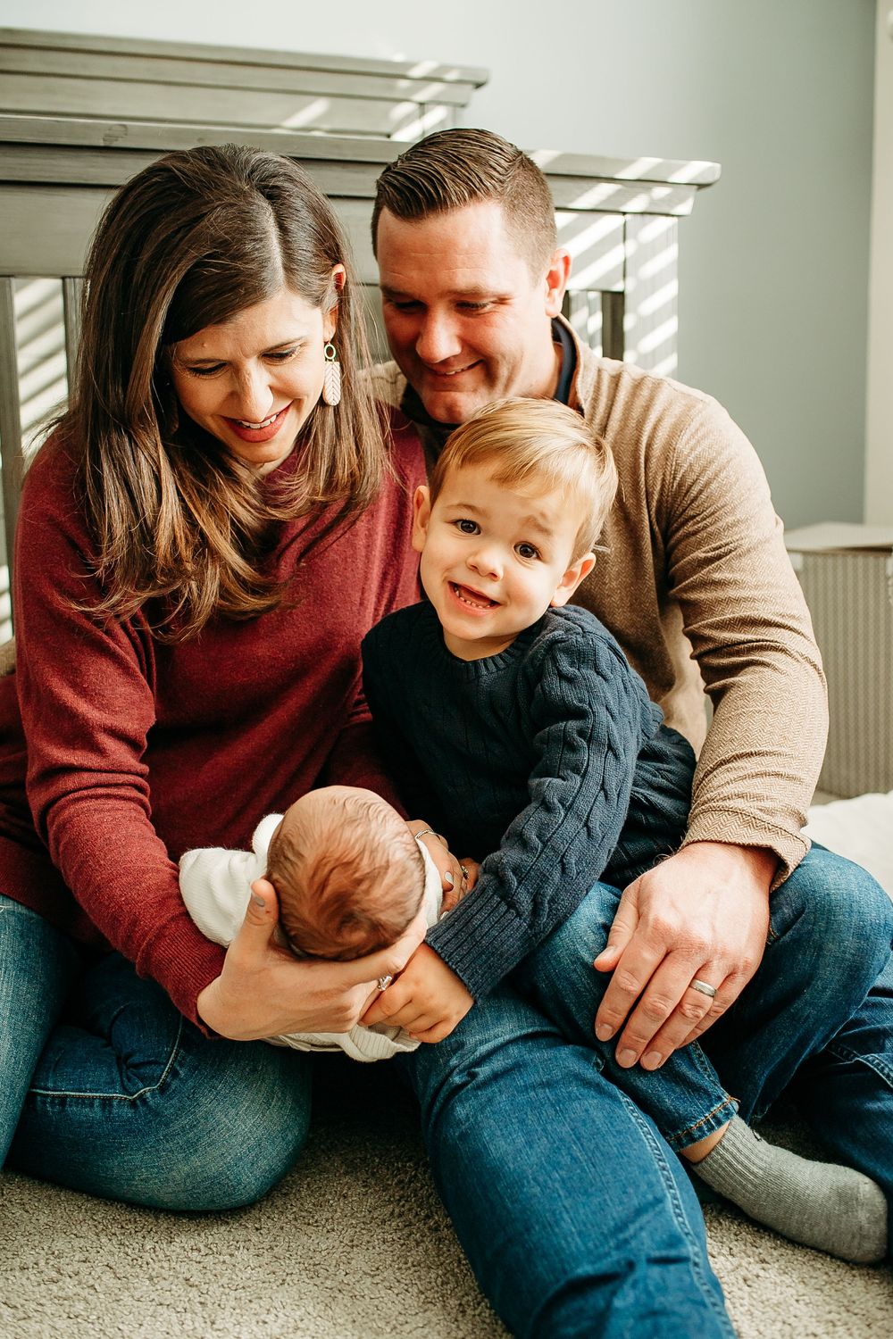Happy family of four sitting together on the floor, parents smiling as toddler holds newborn baby.