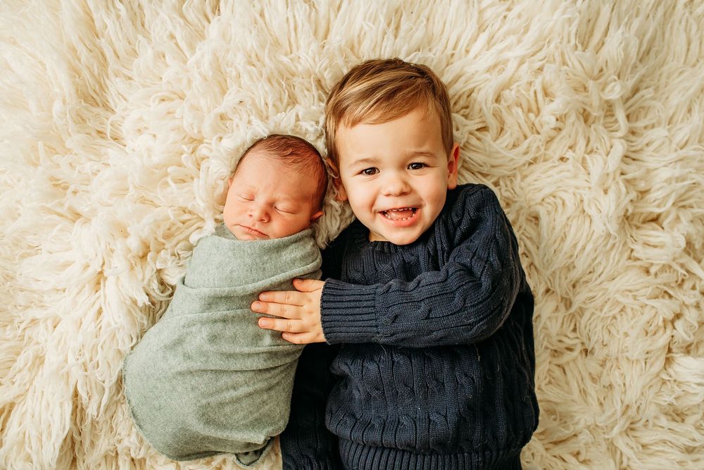 Smiling toddler boy in navy sweater lying beside his swaddled newborn sibling on a fluffy rug.