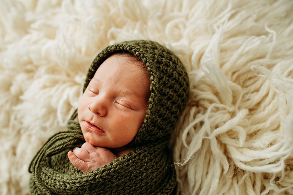 Sleeping newborn baby swaddled in a green knit wrap and bonnet on a cream fluffy backdrop.
