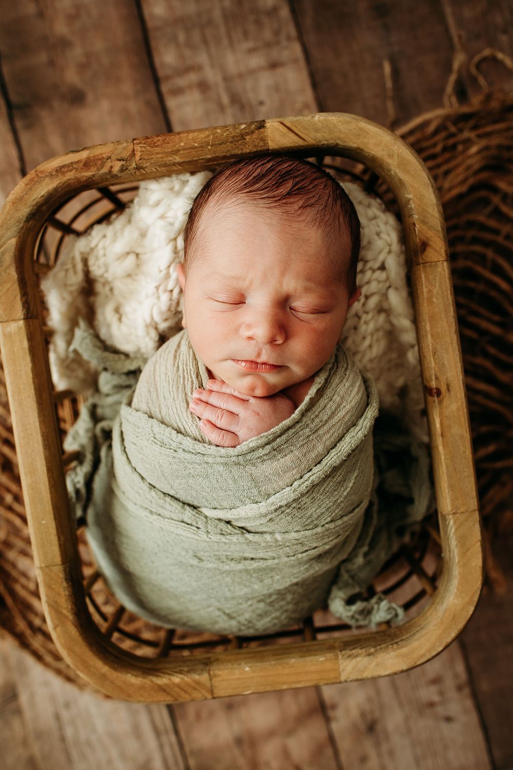 Peacefully sleeping newborn wrapped in sage green swaddle nestled in a rustic wooden basket.