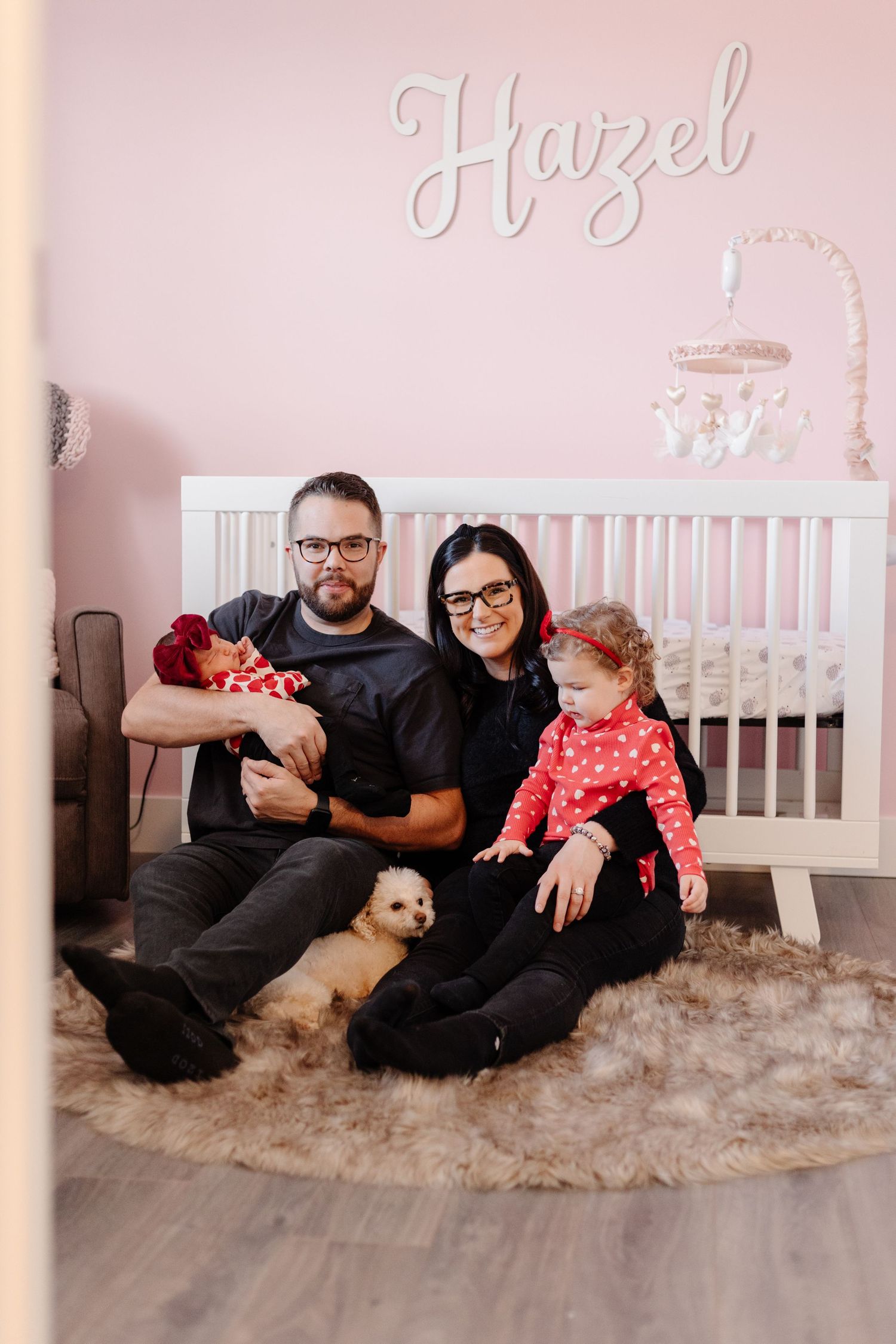 Family with toddler and puppy sitting on fur rug in pink nursery room with Hazel name sign and white crib.